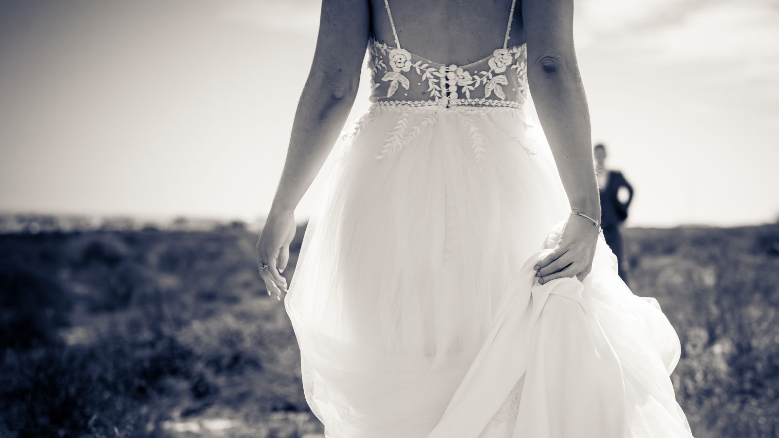 Close-up view of bride's dress and hands, husband in background looking at her.